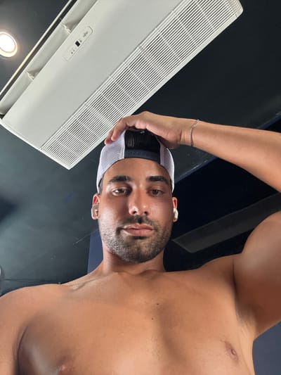 A low-angle, close-up, eye-level, portrait photograph captures a shirtless man wearing a white mesh trucker hat with a dark brim. The man has dark hair, a light beard, and is wearing white wireless earbuds. He is holding his left hand to the top of his head, with his right arm bent at the elbow and raised to his side, flexing his bicep. The background is dark, with the underside of a white air conditioning unit visible at the top of the frame. The man's skin appears to be tanned and slightly sweaty. No specific location in Provincetown, United States of America, can be identified from the image.