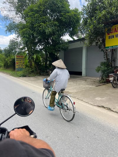 The image depicts a street scene in Thuận Hòa Commune, Vietnam. A person wearing a conical hat and a checkered shirt rides a light blue bicycle on the right side of a paved road. The bicycle has a basket in the front containing blue plastic bags. The rider is wearing green pants and blue shoe covers. In the background, a building with grey roll-up doors is visible, and a motorcycle is parked to the right of the building. A yellow sign with Vietnamese text is mounted on a pole to the left of the road, with some text visible. Trees and foliage line the side of the road. The perspective suggests the image is taken from another vehicle, with a motorcycle's handlebar and a driver's arm visible in the foreground, and a mirror reflecting the driver.