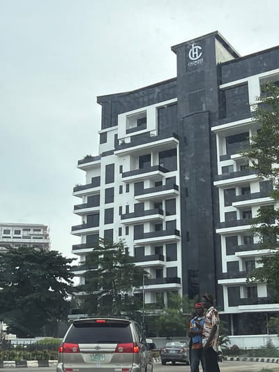 The image shows a partial view of a modern building in Falomo, Nigeria. The building has a distinctive black and white architectural design with projecting rectangular elements. A sign on the building reads "CILONZES Heights." Two people walk past on the sidewalk near traffic. A silver car is partially visible on the right. Trees and a glimpse of another building are also present in the background. The sky is overcast.
