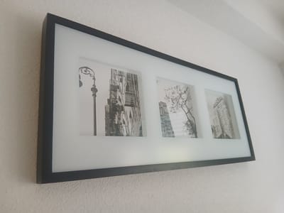 A horizontal, black-framed triptych artwork featuring three black and white photographs is mounted on a lightly textured white wall. The artwork is tilted slightly. The leftmost photograph shows a lamppost with an ornate design and a fire escape on a building. The middle photograph depicts a bare tree branch against a backdrop of buildings. The rightmost photograph features a tall building with signage that reads "GRACE" at the top. The wall has a rough, stucco-like texture. The lighting suggests an indoor environment.