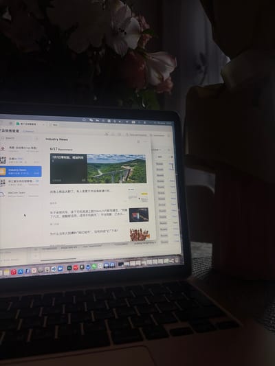 A laptop is open and placed on a dark, textured surface, likely a desk or table. The screen displays a web browser with several tabs open, including one titled "Industry News" featuring articles with images and headlines in Chinese. A dropdown menu labeled "Russia" is partially visible on the right side of the screen. The overall lighting is dim, with light coming from the laptop screen and a shadow from a person's shoulder and head visible in the upper right portion of the image. The environment appears to be indoors, possibly a home office or study. There are no visible signs or indicators suggesting the specific location of Shlisselburg, Russia.