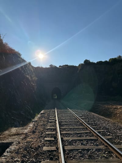 The image depicts a set of railroad tracks leading towards a stone tunnel, nestled between rocky, vegetated embankments under a clear blue sky with the sun shining brightly, causing lens flare. The sun is positioned in the upper left quadrant of the image, casting long shadows. The railroad tracks are made of metal rails supported by wooden sleepers, laid on a bed of ballast. The tunnel entrance is dark and arched, with a faint light visible at the end, suggesting it leads to an open area or another scene beyond. The environment appears to be rural or semi-rural, with dry vegetation and rocks visible on the slopes. The scene suggests an abandoned or disused railway line, given the natural overgrowth and the weathered appearance of the stone structure. The location is identified as Générargues, France. There are no people or animals visible in the image.