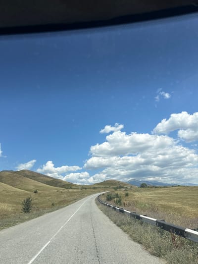 The image shows a winding asphalt road curving through rolling hills covered in dry, golden grass. A black and white guardrail runs alongside the right side of the road. In the distance, mountains are visible under a bright blue sky dotted with fluffy white clouds. The scene appears to be a rural landscape. There are no people or vehicles visible in the image. The location is identified as Рудничный, Kazakhstan.
