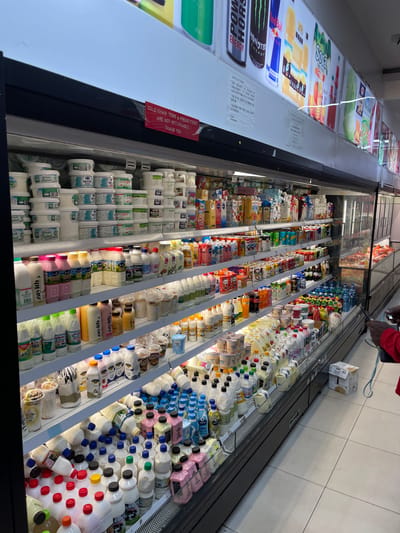 This is a photograph taken inside a well-stocked refrigerated display case in a grocery store located in Guzape, Nigeria. The display case is filled with a wide variety of dairy products, including milk, yogurt, and other beverages, packaged in various bottles, cartons, and tubs. The products are neatly arranged on multiple shelves, with a focus on beverages and yogurts at the bottom. The lighting is bright, illuminating the products and creating a clean, professional appearance. In the background, a person is visible, partially obscured by the display case, holding a smartphone. The overall environment suggests a typical retail setting where perishable goods are kept chilled for sale.