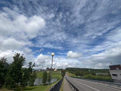 This is a wide-angle, horizontal shot of a road leading into the distance on a cloudy day, likely in the morning or afternoon. The road is paved with white lane markers and is bordered by a dark metal fence. To the left of the road, there is a body of water, lush green grass, and several trees. In the background, rolling hills covered in trees are visible, with a few houses dotted among them. The sky is filled with a dramatic display of white and gray clouds, with patches of blue sky peeking through. The lighting suggests it is daytime, but the heavy cloud cover could be indicative of an approaching storm or just a generally overcast day. There are no people or animals visible in the image. A single, spherical streetlamp stands prominently on the left side of the road, near the foreground. On the right side of the road, towards the background, a building with a red-tiled roof is partially visible, with a small flag on top. The overall mood of the image is serene and natural, with the dramatic sky adding a sense of grandeur to the landscape. The location context provided suggests this scene is from the Kapylsky Selsoviet in Belarus.