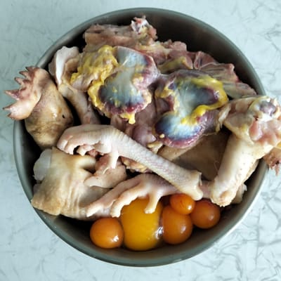 A metal bowl is filled with raw chicken parts, including pieces of chicken feet, chicken gizzards, and what appear to be chicken eggs. The bowl is positioned on a light-colored surface with a marble-like pattern, likely a countertop. The image is taken from a top-down perspective, with the bowl filling most of the frame. The lighting is bright, suggesting an indoor setting. There is no indication of a specific location in Cupcini, Moldova, or any people in the image.