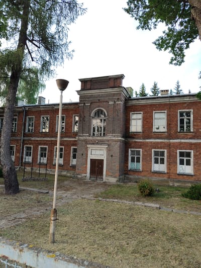 The image shows a two-story brick building with a central entrance. The building has multiple windows with white frames, some of which are barred. The facade is made of red bricks and features decorative brickwork around the windows and the entrance. A tall, narrow chimney is visible on the roof. The building is surrounded by a dry, grassy area with some scattered bushes. A street lamp stands in front of the building, and a large tree is on the left side of the image. The sky is overcast. This building is located at 27 Warszawska Street in Nowy Dwór Mazowiecki, Poland, and is part of the former army barracks complex.