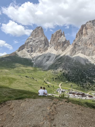A couple, wearing white t-shirts, sits facing away from the camera, embracing each other, on a grassy slope overlooking a valley. In the mid-ground of the valley, a small village with several buildings and a parking lot full of cars is visible. Winding dirt roads lead through the grassy valley, dotted with scattered trees. In the background, majestic, rugged mountain peaks dominate the horizon, their rocky faces catching the sunlight under a partly cloudy sky. The foreground consists of a rocky, gravelly incline. This scene is set in the Dolomites region of Italy, likely near Sëlva - Wolkenstein - Selva di Val Gardena.