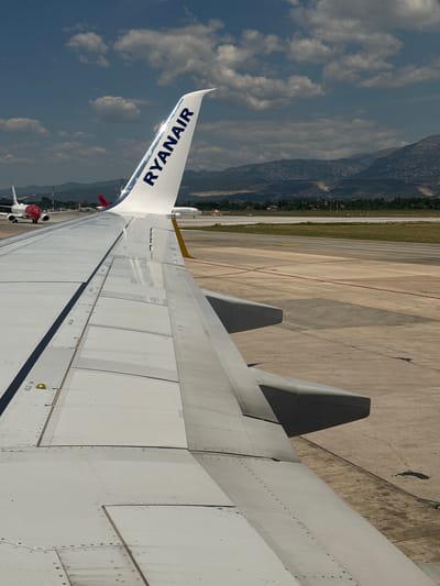 This image captures a close-up view of an airplane wing, likely from the perspective of a passenger seated near the wing, at an airport. The wing is a light grey color with visible panel lines and rivets. A prominent blue "RYANAIR" logo is emblazoned on the wingtip, which is slightly angled upwards. In the background, other aircraft are visible on the tarmac. One aircraft, partially obscured by the foreground wing, is white with a red front. Further in the distance, a runway stretches out, bordered by green grass and trees. Beyond the airport grounds, a range of mountains forms the backdrop under a sky dotted with white clouds. The lighting suggests it is daytime, with sunlight casting shadows on the wing and tarmac. The scene is static and appears to be taken at ground level or during taxiing, not in flight. The location of Kruja, Albania is not depicted or suggested by any elements in the photograph.