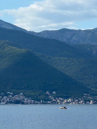 The image shows a landscape in Krašići, Montenegro. The scene includes mountains with varied vegetation, a stretch of coastline with closely arranged buildings, and a body of water reflecting sunlight. In the water, a motorboat is traveling, carrying two people. The sky above the mountains is partly cloudy, with visible blue patches.