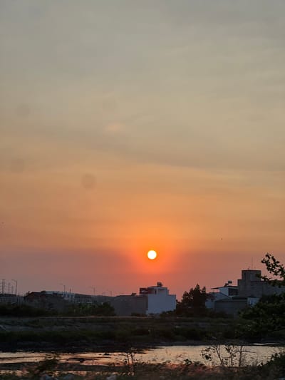 The image depicts a sunset in an urban area, likely in Unknown, Vietnam. The sun is visible as a bright orange orb near the horizon, which is partially obscured by buildings and trees. A body of water runs along the foreground, reflecting the sunset. Buildings, some still under construction, are visible in the background. The sky is filled with a gradient of orange and yellow hues from the setting sun, transitioning to lighter colors at the top of the frame. There are powerlines near the bottom of the image.