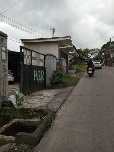 This image depicts an outdoor street scene in Melakudikadu, India, under an overcast sky. In the foreground, a concrete sidewalk runs parallel to a paved asphalt road, with a rectangular concrete drainage ditch containing standing water positioned on the left side of the sidewalk. Adjacent to the ditch, a brown pot holds a plant and a piece of white and striped fabric.
To the left, a dark metal gate, featuring a diamond pattern and yellow-green graffiti spelling "M2R," stands partially open, revealing a white vehicle parked behind it. The gate is supported by concrete pillars. Directly behind the gate and further right is a light-colored single-story building with a sloped roof. An antenna, partially obscured by vine-like vegetation, is visible on the roof. The building's facade includes a dark roll-up door or shutter. Reddish-orange flowering plants grow along the base of the building's fence line.
On the right side of the image, the asphalt road ascends slightly. A person wearing a dark jacket and helmet is riding a dark-colored scooter away from the viewer. Following the scooter is a silver or light-colored SUV. Utility poles and overhead power lines are visible, extending across the upper portion of the frame. The background shows additional residential structures and trees along the ascending road.