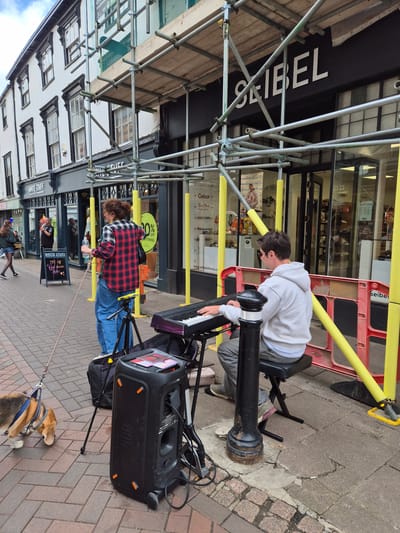 The image depicts an urban street scene in West Suffolk, United Kingdom, featuring two street performers, a dog, and pedestrians, set against a backdrop of commercial buildings partially obscured by scaffolding.

In the mid-ground on the right, a male individual with dark hair and sunglasses, wearing a white hooded sweatshirt and grey trousers, is seated on a black folding stool. He is actively playing a black and purple electronic keyboard, facing away from the camera. To his immediate left and slightly behind him, a female individual with curly brown hair, wearing a red and black plaid shirt and blue jeans, stands. She holds a violin bow in her right hand and a clear plastic water bottle. Her left hand holds a white stick, extending towards the ground.

Between the two performers, a black JBL portable speaker with orange accents and wheels is positioned on the pavement, connected by a power cable. Red-covered items, possibly CDs, are placed on its top surface. Adjacent to the speaker, a small black tripod with a device holder stands. In the lower-left foreground, a small dog, appearing to be a beagle with tan, white, and black fur, wears a blue harness and leash, and is sniffing the brick pavement.

The background consists of multi-story commercial buildings. A significant metal scaffolding structure, fitted with yellow protective padding on its vertical poles, covers the facades of these buildings. Behind the scaffolding, the shop "SEIBEL" (a shoe retailer) is visible with its white lettering on a black background, displaying shoe brands like "Gabor," "Rieker," "Tamaris," and "Josef Seibel" through its windows. To the left of "SEIBEL," the "WHITE STUFF" clothing store is identifiable, with a black A-frame sign on the sidewalk advertising "20% off New season styles." A female pedestrian in a dark top and shorts walks in the background on the left, while another male figure is visible through the "WHITE STUFF" window. A black bollard is present in the foreground, to the left of the seated musician. Red plastic safety barriers are partially visible on the right side of the frame. The pavement consists of red brick pavers and grey concrete slabs, and the scene is illuminated by daylight under what appears to be an overcast sky.