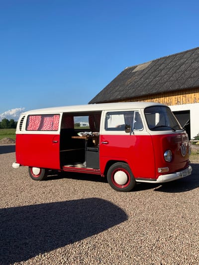 This media file features a vintage red and white Volkswagen Type 2 camper van parked on a gravel surface next to a weathered wooden building. The van is angled, with its driver's side facing the viewer. The building has a dark, textured roof and light yellow, peeling paint on its walls. The van's interior is partially visible, showing red curtains with white polka dots and checkered flooring. There's a person reflected in the driver's side mirror. The sky is a clear, bright blue, indicating daytime with sunny weather. A shadow of the van stretches across the gravel. The license plate on the front of the van reads "CZY 186". The scene appears to be outdoors, possibly in a rural or semi-rural setting, given the building and the presence of trees and distant wind turbines in one of the cropped hints. The overall impression is one of a pleasant, sunny day, highlighting the classic vehicle.