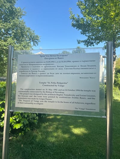 This image presents an outdoor scene featuring a prominent information sign situated in General Todorov, Bulgaria. The sign, constructed of a clear glass pane, is rectangular and vertically oriented, secured by two cylindrical, polished metal posts on its left and right sides. The text on the sign is displayed in both Bulgarian (top half) and English (bottom half).
The sign details the "Temple 'St. Petka Bulgarska'," stating it was "Constructed by Vanga." According to the text, construction commenced on May 31, 1992, and the temple was ceremonially consecrated on October 14, 1994, by the Bishop of Nevrokop. The project was designed by architects Bogdan Tomalevsky and Lozan Lozanov. Academician Svetlin Rusev painted the frescoes and icons, while Grigor Paunov crafted the iconostasis. Vanga's bequest is described as the temple serving as a home for all believers, irrespective of their nationality or religion. The sign concludes with "The 'Vanga' Foundation."
The background of the image features a bright, clear blue sky occupying the upper portion. A large deciduous tree with dense, vibrant green foliage is visible behind and to the sides of the sign, extending into the sky. The tree's leaves are clearly illuminated by natural sunlight. The lower part of the image, below the sign, shows a well-maintained area of lush green grass. Some darker green bushes are also visible near the base of the sign on the left. Faint reflections of a distant building with a visible cross on its roof, presumably the described temple, along with reflections of the blue sky, are discernible on the glossy surface of the glass sign. The overall scene is brightly lit, indicative of a sunny daytime environment.