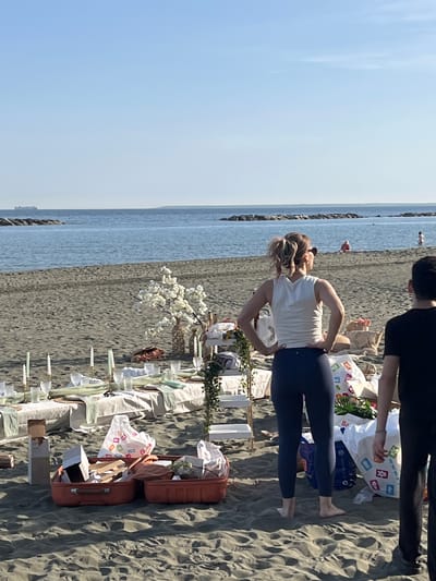 A beach scene in Germasogeia, Cyprus, displays a sparsely populated shoreline with a picnic setup. A woman in a white top and dark leggings stands near a long table laden with items including glassware, candles, and floral arrangements. To the woman's right and slightly above, another individual wearing a black shirt is partially visible. Two open suitcases sit near the table, and bags of goods are strewn on the sand. The ocean occupies the left side of the scene under a clear blue sky.