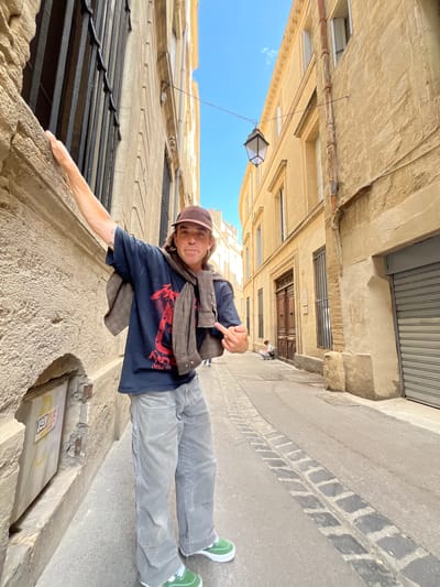 A young man poses for a photo in a narrow street in Montpellier, France. The buildings are old with stone facades. The street surface is paved with stone and asphalt. The man wears a dark T-shirt, gray pants, and green shoes, with a cap and brown jacket around his neck. He extends one arm to touch a wall while pointing with the other hand.