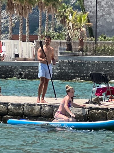 A man stands on a stone walkway at the edge of the water, holding a paddle. He is wearing light blue shorts. In the foreground, a woman is kneeling on a blue paddleboard in the water. She is wearing a bikini. Behind the man are palm trees and some white structures. The water is a vibrant turquoise. The scene appears to be in Risan, Montenegro, a coastal town known for its natural beauty and historical sites.
