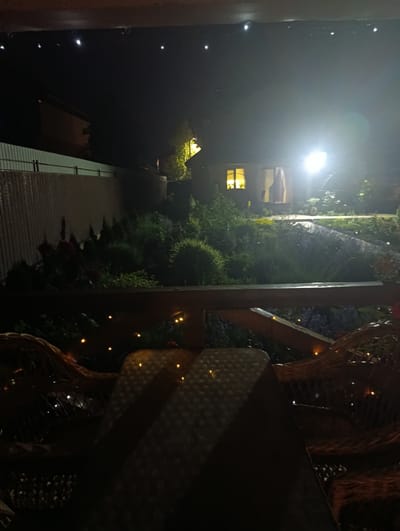 The image depicts a nighttime outdoor scene at a location identified as Korsuntsi, Ukraine. The foreground features a wooden railing and wicker furniture illuminated by small string lights. Beyond the railing, a dimly lit garden area is visible with various plants and bushes. In the background, a house or building is present, with a window emitting a warm yellow light and another area brightly illuminated by a strong external light source, casting a glow on the surrounding garden and a path. The sky is dark, with a few small, bright lights visible against it, possibly decorative string lights. The overall atmosphere is one of a peaceful, illuminated evening in a residential garden.
