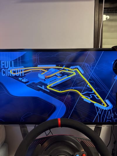 A monitor displays a racing circuit map in blue and yellow. The track is outlined in yellow, with starting grid and pit lane details visible. A racing wheel is partially visible on the right edge of the image, with a black rim and a red stripe. The "FULL CIRCUIT" text is visible at the bottom, indicating the circuit layout being shown. A white wall section is visible to the left.