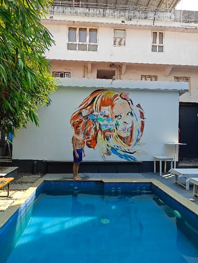 An outdoor scene in Kochi, India, features a fair-skinned male in blue swimming trunks standing barefoot at the edge of a rectangular swimming pool filled with clear blue water. He faces forward, shirtless, with arms at his sides. Behind him, a large, vibrant mural depicting two abstract, intertwined female faces in an expressive style with orange, red, blue, green, and white hues covers a white wall.
Above the mural wall, a multi-story white building with several framed windows and an open balcony structure is visible. To the left of the pool, lush green foliage, resembling bamboo, partially frames the scene. To the right of the pool, white outdoor furniture consisting of two small tables with chairs and two lounge chairs is present on a paved area. The environment suggests a recreational space within a residential or hospitality complex.