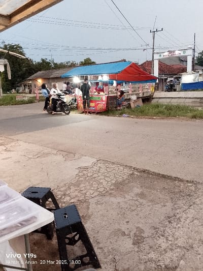 The image shows a street food stall with a red and blue awning in Kecamatan Kroya, Indonesia, at latitude -6.422 and longitude 107.996. The stall is on the side of a paved road. There are a few people standing around the stall, including a person wearing a blue shirt. In the background, a sign arch that says "DIBERIKAN JAYA JAYA" is visible. The street is empty, with only a single motorcycle parked near the stall. Two black folding chairs are in the foreground. The sky is overcast and the lighting is dim, indicating it is evening.