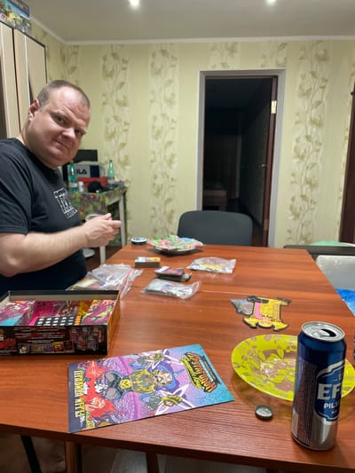 A medium shot shows a man in a black t-shirt sitting at a wooden table, engaged in a board game. The man's left arm is bent, and his hands are near a clear plastic bag containing game cards. A colorful board game box, partially opened, is visible to the left of the man. Several other game-related items, including cards in plastic sleeves and a decorative game piece, are scattered on the table. A can of Efes Pilsen beer, an empty yellow plate with some residue, and a bottle cap are also present. The background features a doorway leading to another room and wallpaper with a floral pattern. The scene appears to be in a domestic setting in Novocherkassk, Russia.