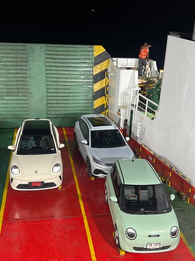This media file shows three cars parked on the deck of a ferry at night. The cars are a light green mini, a white car, and a light cream colored car. A person wearing an orange vest is standing on the ferry near the cars. The ferry appears to be docked, as suggested by the presence of a large green container in the background. The ground of the ferry is red with yellow and black striped markings. The sky is dark, indicating it is nighttime. The location is Nathon, Thailand. There is text visible on the license plates of the cars and on signs on the ferry, but it is difficult to read clearly.