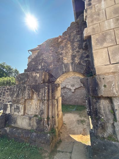 The image shows the exterior of the Romanesque former monastery church of Walkenried Abbey, located in Walkenried, Germany. The sun is bright and directly overhead, casting a strong glare in the upper left corner. The focus is on a weathered, arched stone entryway that leads into a courtyard. The stonework is ancient and shows signs of age and erosion, with moss and lichen growing on the surfaces. To the right of the arch, a wall made of lighter, cut stone blocks is visible. To the left, there is a stepped stone structure, also weathered and partially overgrown. In the background, through the arch, the courtyard appears to be a mix of dirt and grass. Trees and part of a building with a tiled roof are visible in the upper left quadrant. The scene suggests a historical site that is partially in ruins but maintained. There are no people visible in the image.