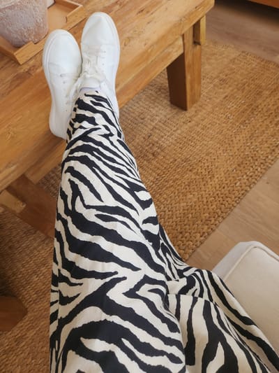 The image shows a person sitting with their legs crossed, wearing white sneakers and black and white zebra-print pants. The person is seated on a woven rug, with a wooden surface visible beneath their legs. A portion of a cream-colored chair or sofa is also visible. The setting appears to be indoors, possibly a home or studio, with natural light illuminating the scene. The focus is on the person's attire and the textures of the surrounding elements. There is no visible text in the image.
