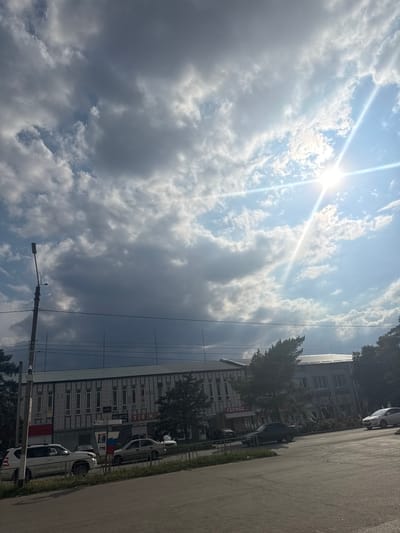 This image, taken in Yegorlykskaya, Russia, captures a street scene under a partly cloudy sky with the sun prominently visible and emitting strong rays. The sun's glare suggests it is daytime, likely late morning or early afternoon. The sky is a mix of bright blue with scattered white and grey clouds, indicating variable weather.

The scene features a paved road on the right side of the frame, with a grassy strip and a low fence separating it from a building. The building appears to be a multi-story structure, possibly a residential or commercial building, with a modern facade featuring many windows and decorative elements. Several cars are parked along the building or on the street. Trees line the area in front of the building, partially obscuring it in some sections. A utility pole with wires and a street lamp is also visible, typical of an urban or suburban environment.

There is no discernible activity or event taking place, suggesting a still moment in time. The overall mood is neutral, with the bright sunlight contrasting with some of the darker clouds, creating a dynamic sky. Visible text on the building includes "ГЕРМЕС" and "ОСАГО". The image is oriented with the sky at the top left and the road and building at the bottom right, suggesting the photo was taken from a low angle or rotated.