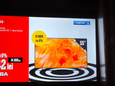 The image displays a television advertisement. The advertisement features a large, vibrant screen showcasing a television with an abstract, fiery orange and yellow image displayed on it. The television is identified as "Neo QLED" and is 55 inches in size. A graphic overlay indicates "CINEMA 1" in a blue rectangular box.
To the left of the television screen, there is a red section with text. This text appears to be promotional, detailing a sale or offer. Visible text includes "12 RATE cu 0%", suggesting 12 installments with 0% interest. Below this, the number "42" is prominently displayed in large font, followed by "lei" and "BA". Another black box shows "14 899 lei". The red section also includes partial text related to "samsung" and "UXUA 55" / Neo QLED".
The overall scene suggests a retail or promotional display, likely in a store or as part of a television broadcast. The lighting is bright, focused on the advertisement. No people are visible in the image. The background of the main advertisement screen is white, with a black and white circular pattern beneath the television, possibly representing sound waves or a stylized base. The mood is promotional and informative, highlighting the product and its payment options. The location context of Ocnița, Moldova, is provided externally.