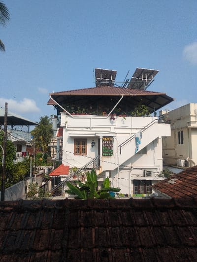 The image depicts a residential scene in Kochi, India, under a clear blue sky. In the foreground, a dark, weathered, traditional tiled roof is partially visible, indicating the viewer's elevated perspective. The central subject is a multi-story, white-painted building with architectural details. Its upper level features a brown hip roof with a flat section where a rooftop garden or planted area is present. Above this, two distinct arrays of solar panels are mounted on metal frameworks, angled for solar exposure.
An external white concrete staircase ascends the right side of the building, leading to the roof level; a blue and white patterned cloth is draped over its railing. The facade includes a brown wooden-framed window and a green-framed window with security bars. Below the brown window, a green and yellow sign with Malayalam script is affixed. Potted plants are visible on the balconies and ledges. A white air conditioning condenser unit is mounted on the exterior wall to the right.
The mid-ground features dense tropical foliage, including banana plants, partially obscuring the lower sections of the main building. In the background, a tightly packed urban environment is visible with various residential structures, some featuring red-tiled roofs and white walls. Utility poles and a network of overhead wires are present. Palm trees and other greenery are scattered throughout the scene. No individuals or specific human actions are discernible.