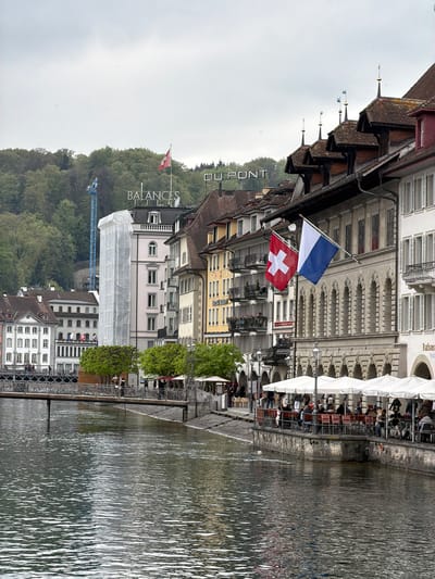 A detailed view captures a cityscape in Lucerne, Switzerland, along a canal. Traditional buildings line the waterfront, adorned with Swiss and Lucerne flags. One building features an outdoor restaurant with seated patrons. A green hillside looms in the background, partially obscured by building structures. A construction crane rises above the landscape. The canal's water surface reflects the buildings and sky. The image is oriented vertically, showing details in the architecture and the bustling life of the city's waterfront.