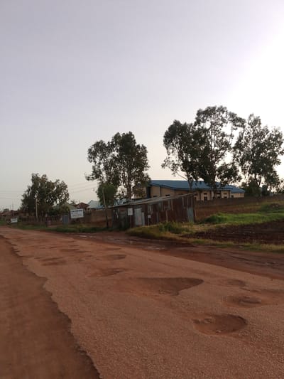 The image depicts a rural road in Barakin Angundi, Nigeria, characterized by its rough and uneven surface with numerous potholes filled with water. The road appears to be made of dirt or gravel. On the left side of the road, there are some buildings and trees. One building has a sign that reads "BORNO GOOD NEWS". Further down the road, there is a larger building with a light blue roof, possibly a school or church, surrounded by a wall. On the right side of the road, there is a dilapidated structure made of corrugated metal, and beyond it, a field of crops. The sky is overcast, suggesting a cloudy day. The overall scene portrays a somewhat underdeveloped area with basic infrastructure.