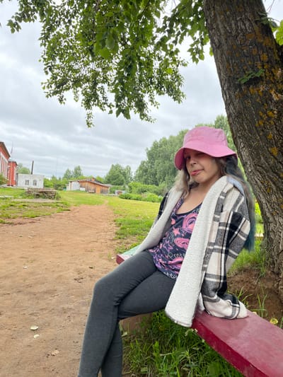 In the village of Baldeika, Russia, a young girl wearing a pink bucket hat, a patterned tank top, a plaid jacket, and dark leggings sits on a red bench next to a large tree. Her left leg is crossed over her right, and she is sticking out her tongue, looking towards the camera. The background consists of a dirt path, green grass, trees, and several small buildings. The sky is overcast.