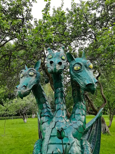 The image shows a large, green, metallic sculpture of a three-headed dragon situated in a park. The dragon sculpture has detailed scales and bright yellow eyes. It is positioned in front of a backdrop of lush green trees with thick foliage. The ground is covered in green grass, and there are several other trees visible in the background, some with twisted trunks. The sky is overcast. This sculpture is located in Zelenogorsk, Russia.