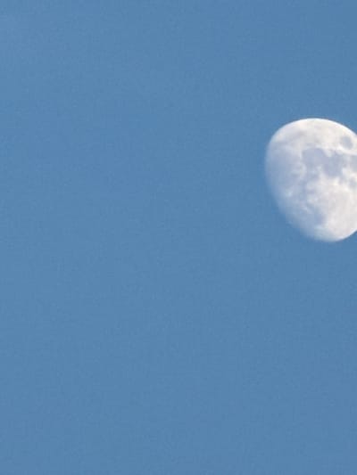 A partially illuminated moon is visible against a clear blue sky. The moon, which is in its waxing gibbous phase, is positioned in the upper right quadrant of the image. Its surface shows visible craters and shadowed areas, indicating it is illuminated by sunlight. The sky is a uniform, uninterrupted expanse of blue, suggesting it is daytime or early evening. The image provides no information about the location within Riverhead, United States of America, or any other contextual details.