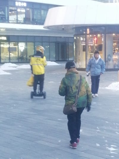 An outdoor pedestrian plaza in Fengtai District, China, is depicted under diffused daylight, with scattered snow patches on a light-colored planked surface. Three individuals are present. In the foreground, a person walks away from the camera, wearing a dark cap, a camouflage-patterned padded jacket, dark trousers, red sneakers, and a brown cross-body bag. In the middle ground, an individual, likely a delivery worker, operates a black self-balancing scooter. They wear a yellow helmet, a bright yellow jacket, dark trousers, and carry a yellow bag. In the background, a male individual walks, dressed in a light blue puffer jacket over a white hoodie, dark blue trousers, and white sneakers. Modern commercial buildings with extensive glass facades and some illuminated storefronts are visible behind the individuals, with snow accumulated at their foundations.