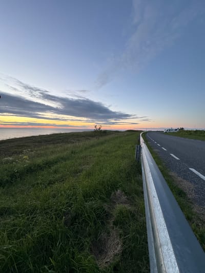 This image depicts a coastal road in Paldiski, Estonia, at dusk. A paved road curves to the right, bordered by a metallic guardrail on the left. Tall green grass and some wildflowers grow along the roadside. In the distance, a white caravan or RV is parked on the right side of the road. The sky is a gradient of blue, transitioning to soft hues of orange and yellow near the horizon where the sea is visible. Wispy clouds stretch across the sky. The overall scene suggests a tranquil evening setting in a rural coastal area.