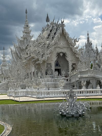 The image shows the Wat Rong Khun, also known as the White Temple, a Buddhist temple in the Chiang Rai province of Thailand, located in the district of Mueang Chiang Rai, Ban Rong Khun. The temple is an ornate white structure adorned with intricate carvings and sculptures. It is surrounded by a moat and lush green grass. In the foreground, a white fountain-like sculpture sits in the water. The sky above is cloudy with patches of blue. Two individuals are visible standing on a balcony of the temple. The overall scene conveys a sense of architectural grandeur and artistic detail within a serene environment.