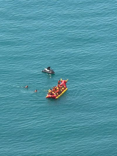 An aerial, wide shot shows a group of people in the sea off the coast of Dzhankhot, Russia. In the foreground, a bright red inflatable boat shaped like a bull is filled with numerous people wearing yellow life vests. To the left of the inflatable, three individuals are swimming in the water, each wearing an orange-red life vest. Further to the left and slightly behind the swimmers, a black and white jet ski is being operated by a person. The water is a consistent blue-green color with moderate ripples. The scene appears to be a recreational activity on a sunny day.