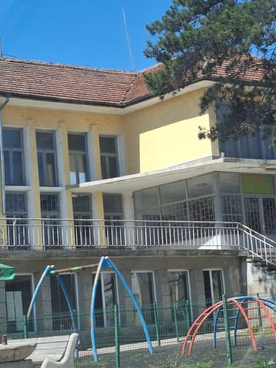 The image shows a building in Studeno Buche, Bulgaria. The building is multi-story with windows and a fire escape. There is a tiled roof on one section of the building and a yellow facade on another. A playground with colorful playground equipment is visible in front of the building. A tree is partially obscuring a portion of the building. The sky is blue.