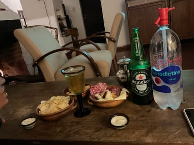 In El Rodeo, Argentina, a close-up shot captures a dimly lit wooden table adorned with food and beverages. A glass of amber liquid, likely beer, sits beside a bowl of potato chips and a small dish of white dip. Another bowl contains a charcuterie assortment, featuring sliced salami and pieces of bread, accompanied by a slice of cheese. To the right, a green bottle of Heineken beer stands next to a large, clear plastic bottle of Benedectino sparkling water with a red spray nozzle. In the background, two upholstered chairs with wooden frames are visible, along with a rustic wooden cabinet and shelves displaying various items. The overall ambiance suggests a relaxed evening gathering.