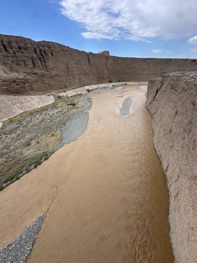 The image depicts a wide, muddy river flowing through a canyon in Jiayuguan, China. The canyon walls are steep and eroded, with layers of brown soil and rock visible. The river is a light brown color, indicating it is carrying a significant amount of sediment. On the left bank, there is a constructed embankment made of concrete blocks and gravel, with some sparse vegetation. On the right bank, the canyon wall is closer to the river and appears to be made of the same eroded rock. The sky above is blue with scattered white clouds. The scene is natural and appears to be a remote area, possibly a geological park or a protected natural reserve within Jiayuguan. There are no people or man-made structures immediately apparent in the river or canyon, except for the embankment.