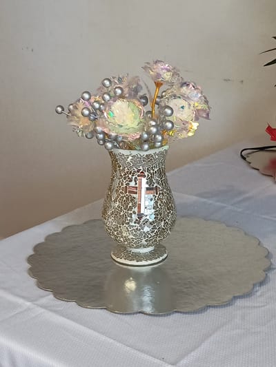 A mosaic vase filled with iridescent flowers and silver beads sits on a scalloped silver platter. The vase is decorated with a mosaic pattern and features a cross made of brown and beige tiles. The platter is placed on a white tablecloth. The background is a plain, light-colored wall. The image appears to be taken indoors, possibly in a home or a place of worship. There is no indication of the specific location within Amarillo, United States of America.