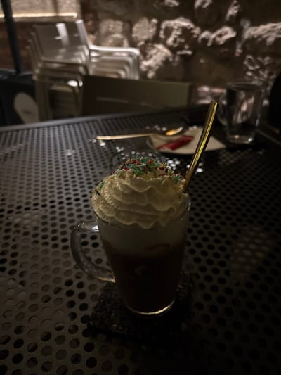 A low-light, portrait-oriented photograph depicts a cold beverage in a clear glass mug, topped with a generous swirl of whipped cream and colorful sprinkles. A golden spoon is inserted into the drink. The glass mug features a handle on its right side and sits on a dark, perforated metal table or tray, which occupies the majority of the foreground and midground. The drink itself appears dark brown, possibly chocolate or coffee-based.

In the blurred background, to the left of the main subject, a rustic, textured stone wall is visible, suggesting an older building. Below the stone wall, several light-colored, possibly white or grey, chairs are stacked or arranged, further indicating an indoor setting like a cafe or restaurant. The overall lighting is dim, suggesting an evening setting or a cozy, subdued ambiance. There are no people visible in the image, and no discernible activity beyond the presence of the drink. No text is visible. The location context indicates this scene is in Alba Iulia, Romania.
