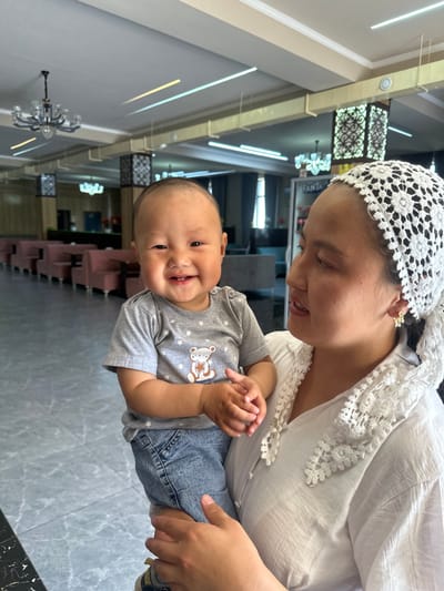 The image depicts a woman holding a smiling baby in an indoor setting that appears to be a lobby or lounge area, possibly in Ынтымак, Kyrgyzstan. The woman, wearing a white, lacy headscarf and a white top with lace trim around the collar, is looking down at the baby with a gentle expression. She has a gold floral earring in her visible ear. The baby, dressed in a gray t-shirt with a bear and stars design and denim jeans, is looking directly at the camera with a wide, toothy grin. The background shows a long row of cushioned seating in a muted pink color, tables, and decorative lighting fixtures on the ceiling, suggesting a public or commercial space. The flooring is a tiled surface.
