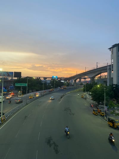 The image captures an elevated view of a multi-lane road and an elevated train line at dusk in St. Thomas Mount-Pallavaram Cantonment Board, India. The sky displays a gradient of soft blues and warm yellows, indicative of sunset. The road is moderately trafficked with several cars, motorcycles, and auto-rickshaws. On the right side of the road, a row of parked yellow auto-rickshaws is visible. A modern concrete elevated train track runs above the road, with a train station structure visible in the distance. On the left side of the road, there are billboards and a building with multiple floors. Lush green trees line parts of the roadside. The scene suggests an urban environment during the transitional period between day and night, with artificial lighting starting to illuminate the road.