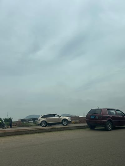 The image shows a roadside scene under an overcast sky. The primary subjects are two parked cars: a dark red hatchback and a lighter colored SUV or crossover. The red car is parked closer to the foreground, and the lighter vehicle is behind it. Both cars are parked along the side of a paved road.
In the background, there are some structures that appear to be under construction or are industrial in nature. Further in the distance, hills or mountains are visible. The ground beside the road appears to be a mix of dirt and some vegetation, with a concrete curb separating it from the road.
The weather appears to be cloudy, as indicated by the uniformly gray sky. The lighting suggests it could be daytime, but the overcast conditions prevent strong shadows.
There is a person standing near the front of the lighter colored car, and further down the road, two motorcycles are parked, with one person near them. This suggests a potential rest stop or a gathering place.
No discernible text is visible on the cars or in the surrounding environment. The overall impression is of a quiet roadside scene in a developing or rural area. The setting could be described as a peri-urban or rural roadside, possibly in Crescent, Nigeria, given the context. There are no strong emotional cues conveyed by the image; it is a straightforward depiction of the scene.